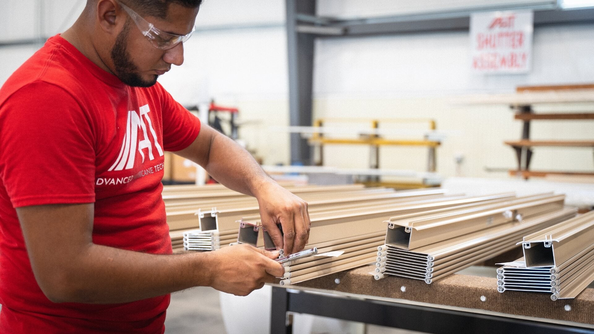 An AHT team member in safety glasses assembling roll-down shutter components at a workbench in AHT's factory