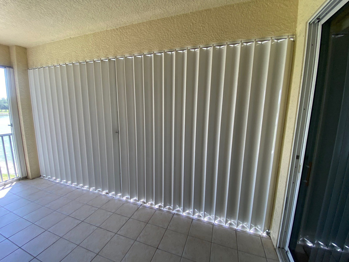 White accordion shutters closed across a finished balcony opening, with center locks and top and bottom tracks.
