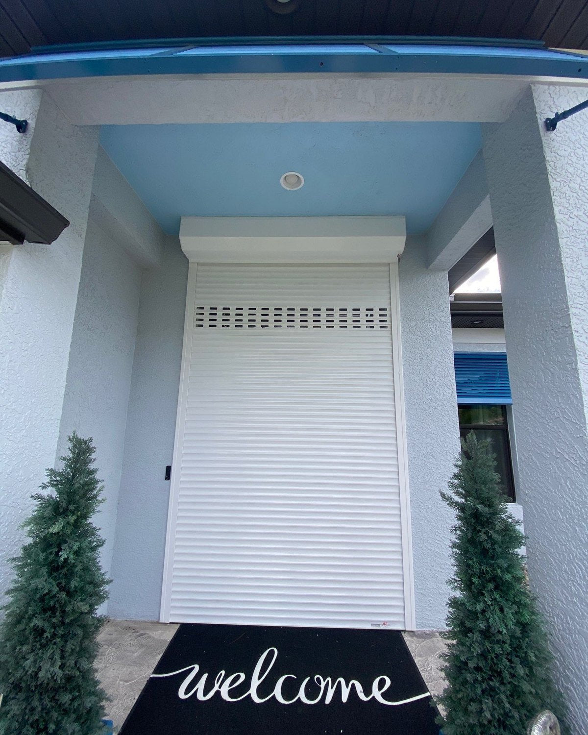 White roll-down shutter closed over a front door, potted evergreens on each side
